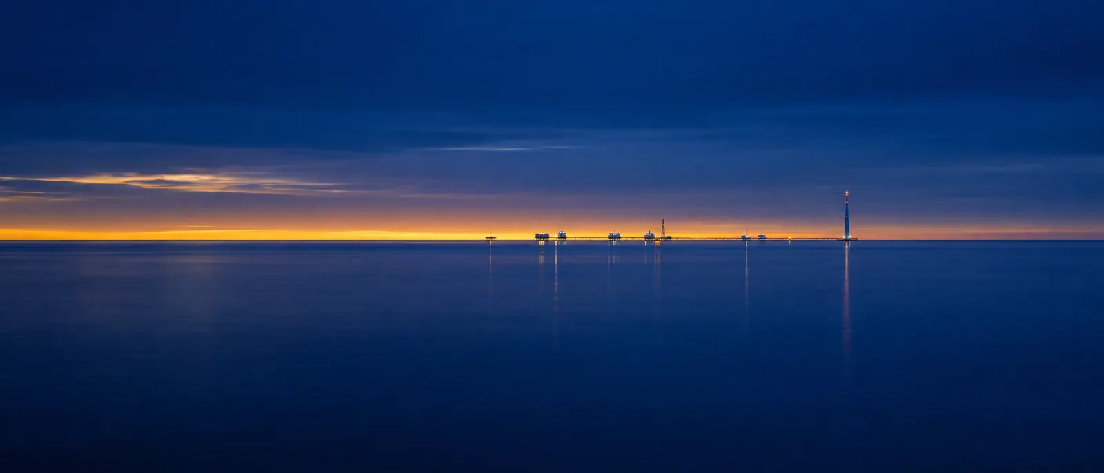 A calm navy ocean at twilight with a thin gold horizon and distant lights